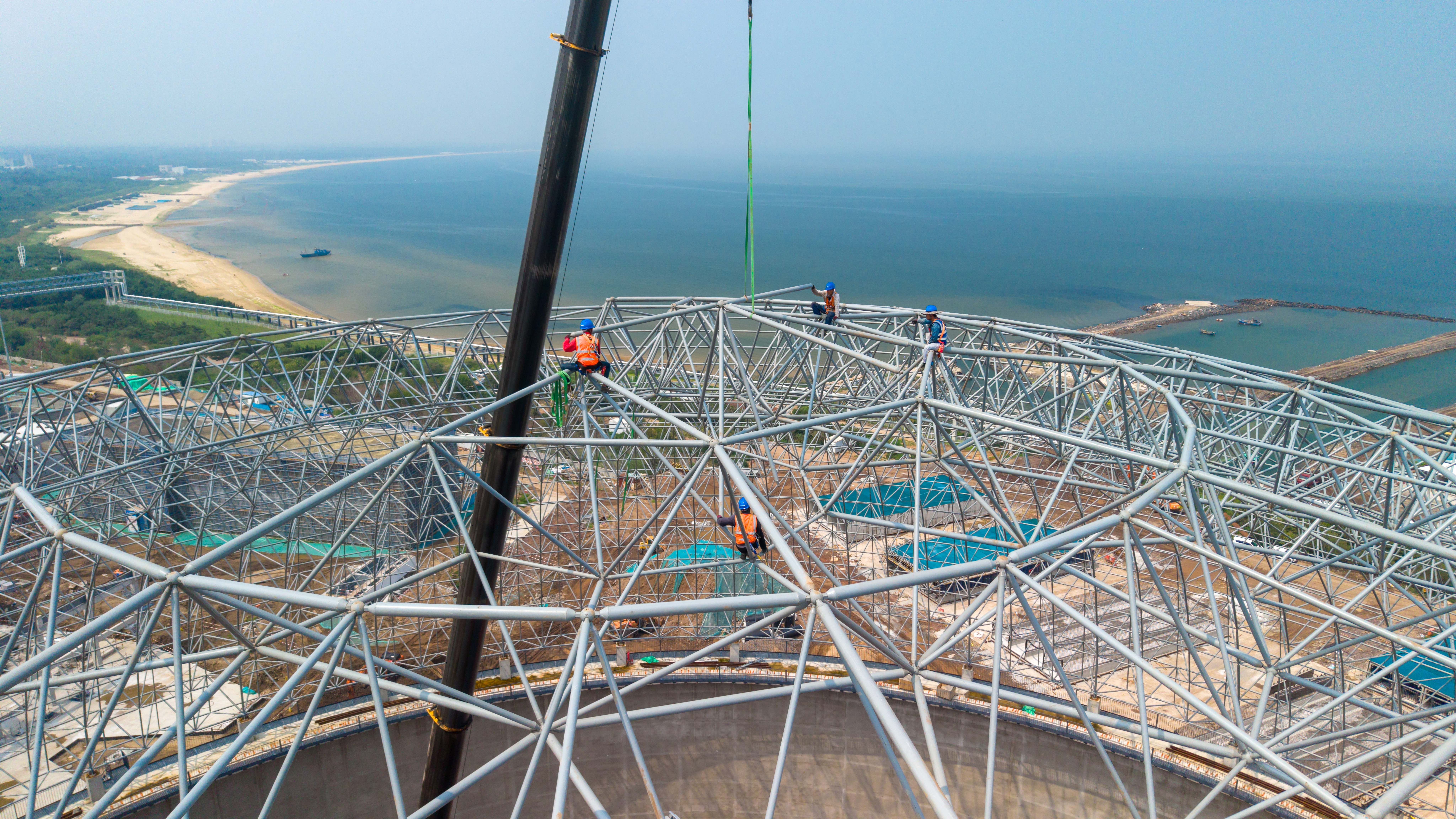 A group of adult male construction workers in safety gear work on a large curved steel dome framework at a coastal construction site. The elevated view shows a sandy beach, calm sea, and clear sky in the background, with construction equipment and infrastructure development visible on-site.